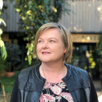 Woman in black jacket looking away outdoors with greenery and building behind
